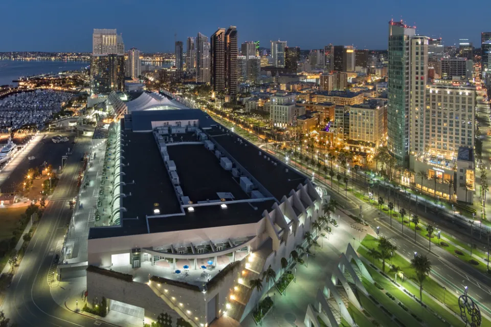 The San Diego Convention Center is illuminated at night with the backdrop of public transit lines and high-rise buildings.